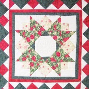 May include: A square quilt featuring a central star pattern composed of red rose floral fabric and cream-colored patterned fabric. The design includes a white octagonal center, a red border, and an outer edge of dark green and red diamond shapes.