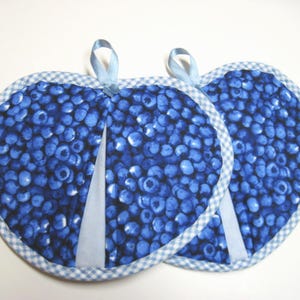 May include: Two heart-shaped pot holders with a blue blueberry pattern. Each pot holder has a light blue gingham trim and a light blue ribbon loop for hanging. The background is white.