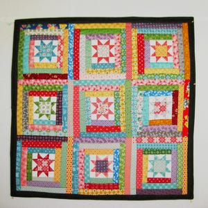 May include: A square patchwork quilt featuring nine blocks arranged in a grid. Each block contains a central eight-pointed star surrounded by concentric fabric strips in various colors and patterns, all framed by a solid black border.