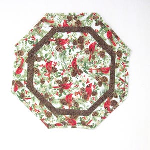 May include: Two octagonal placemats with a festive winter design. The placemats feature a white background with red cardinals, pinecones, and holly. A brown patterned border frames the inner octagon. The placemats are likely made of fabric.