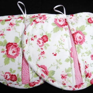 May include: Two heart-shaped pot holders with a white background and a floral pattern of pink and red roses and green leaves. The pot holders have a pink polka dot accent and a white ribbon loop for hanging.