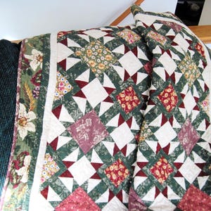May include: A handmade quilt with a star pattern in shades of green, white, and burgundy. The quilt features floral and geometric patterns, with a white border and a green floral edge. The quilt is draped over a dark green couch.