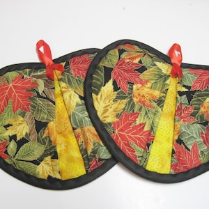 May include: Two quilted, heart-shaped potholders. The potholders feature a black border and a colorful leaf pattern in red, green, and yellow. A yellow fabric strip with a red ribbon loop is attached to each potholder.
