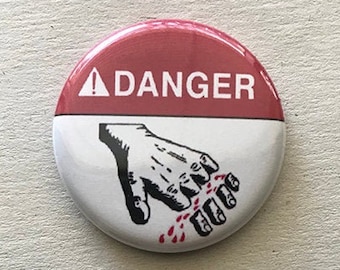 May include: A red and white button with a black and white illustration of a hand with bloody fingers. The button reads "DANGER" with a black triangle and exclamation point.