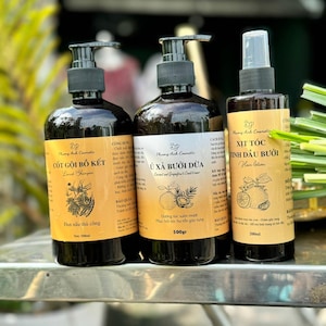 May include: Three brown bottles of hair care products with labels in Vietnamese. The bottles are labeled "Cót Gội Bồ Két", "Ủ Xả Bưởi Dừa", and "Xịt Tóc Tinh Dầu Bưởi". The bottles are on a silver surface.