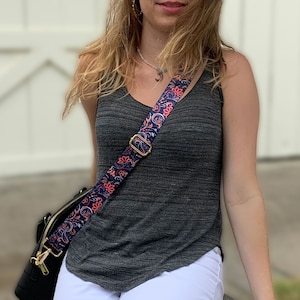 Puede incluir: Una mujer con un top gris sin mangas y pantalones blancos lleva un bolso negro con una correa estampada de colores. La correa tiene un diseño de paisley con colores rosa, azul y naranja.