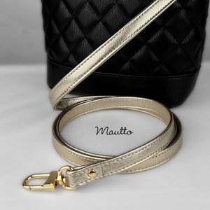 May include: A black quilted bucket bag with a gold chain strap and a gold shoulder strap. The strap is detached and laid out on a white surface. The bag has the word "Mautio" printed on the strap.