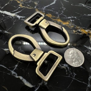 May include: Two antique brass-colored swivel clasps with rectangular and oval loops. A silver quarter coin is next to the clasps for size comparison. The clasps are on a black and gold marble surface.
