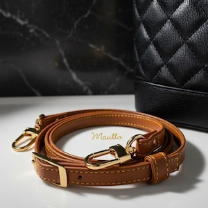 tan vachetta leather strap with yellow (gold) stitching, buckle adjuster and crossbody length. Shown next to a black bag.