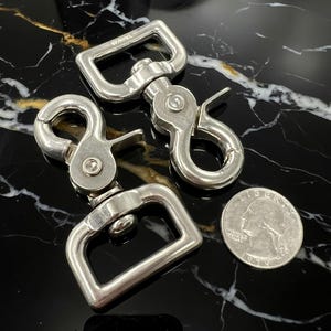 May include: Three silver-colored swivel snap hooks with rectangular loops and spring-loaded clasps. A US quarter coin is included for size comparison. The hooks are displayed on a black, marble-like surface.