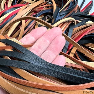 May include: A collection of thin, flat leather strips in various colors including black, tan, light beige, and red. The strips are arranged in a tangled, overlapping pile, showcasing the textured surface and flexible nature of the leather material.