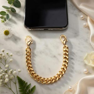 May include: A gold-colored chain phone strap with lobster claw clasps. The chain has a thick, curb link design. The phone is black and has a clear case. The background includes white flowers, greenery, and a white satin cloth.