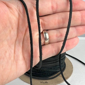 May include: A spool of black leather cord, approximately 3 mm wide, is shown in the image. The cord is wrapped around a cardboard spool and is being held in a hand. The text "Mauitto" is printed on the spool.