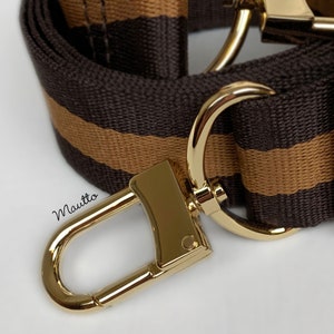 May include: A brown and tan striped fabric strap with a gold metal hook and loop closure. The strap is designed for a bag or purse.