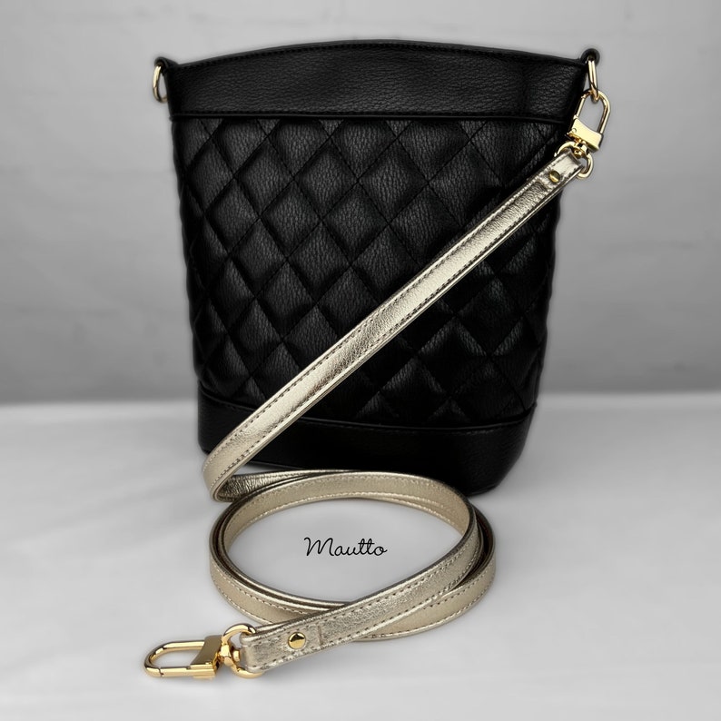 May include: A black quilted bucket bag with a gold chain strap and a gold shoulder strap. The strap is detached and laid out on a white surface. The bag has the word "Mautio" printed on the strap.