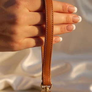 Dark Tan leather wrist strap with yellow stitching and gold clip, held in a hand.