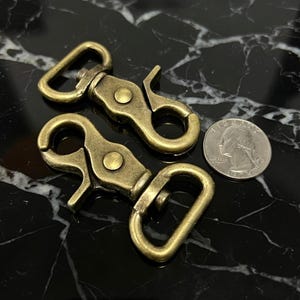 May include: Two antique brass-colored swivel snap hooks with a quarter coin for size comparison. The hooks have a spring-loaded clasp and a swivel eye. The metal has a slightly aged appearance.