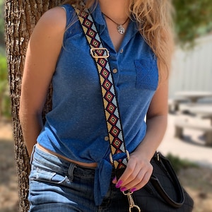 May include: A woman wearing a blue shirt tied at the waist and jeans is holding a black handbag with a patterned strap. The strap is black, red, yellow, and white with geometric designs.