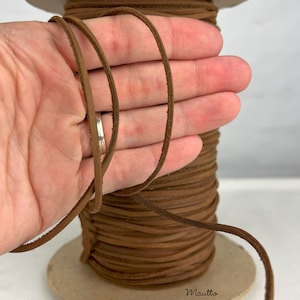 Puede incluir: Un carrete de cordón de cuero marrón, de aproximadamente 3 mm de ancho, está envuelto alrededor de un carrete de cartón. El cordón se sostiene en la mano de una persona para mostrar el ancho y la textura.