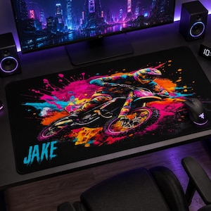 May include: A rectangular desk mat featuring a vibrant, multi-colored graphic of a person riding a motocross bike. The design includes splashes of pink, orange, and blue paint. The name JAKE is printed in a light blue, stylized font in the corner.
