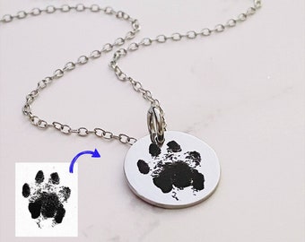 May include: A silver-toned circular pendant necklace featuring a black engraved animal paw print. The pendant is attached to a delicate silver chain. A small square inset shows the original black paw print graphic with a blue arrow pointing to the pendant.