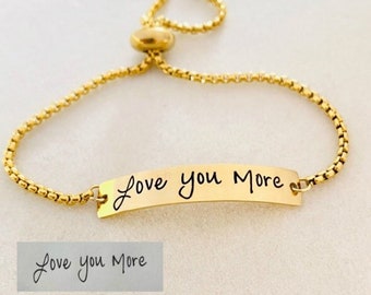 May include: A gold-toned adjustable slider bracelet featuring a curved metal bar engraved with the black script text "Love you More". The bracelet has a delicate box chain design with two small spherical beads at the ends of the sliding mechanism.