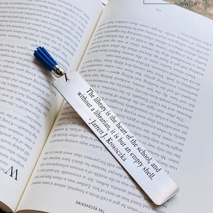 Personalized stainless steel engraved bookmarks with custom text and tassel accents displayed in an open book. Laser engraved metal bookmarks feature inspirational quote and customizable fonts, creating durable personalized gifts for readers, teachers, graduates, and book lovers.