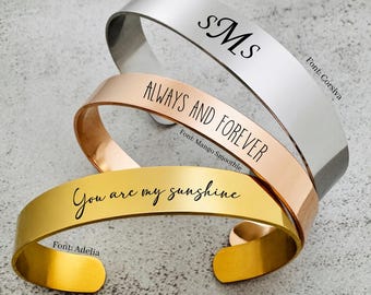 May include: Three metal cuff bracelets in silver, rose gold, and gold tones. The silver bracelet features the monogram "sMs" in Corsiva font. The rose gold bracelet is engraved with "ALWAYS AND FOREVER" in Mango Smoothie font. The gold bracelet reads "You are my sunshine" in Adelia font.