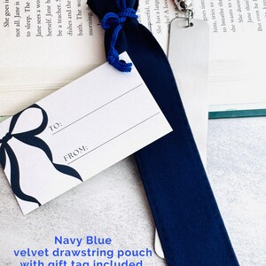 May include: A navy blue velvet drawstring pouch with a matching blue cord, accompanied by a white gift tag featuring a black bow illustration and the printed text TO: and FROM:. A silver metal bookmark with a pink tassel is partially inside.