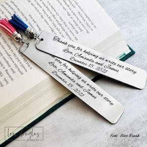 May include: Two rectangular silver metal bookmarks with rounded ends, each featuring a pink or blue tassel attached by a silver ring. The bookmarks are engraved with the text: Thank you for helping us write our story, Love, Amanda and James, December 15, 2021.
