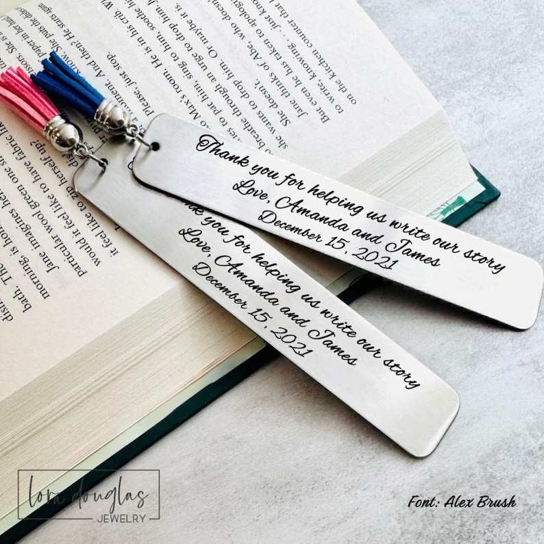 May include: Two rectangular silver metal bookmarks with rounded ends, each featuring a pink or blue tassel attached by a silver ring. The bookmarks are engraved with the text: Thank you for helping us write our story, Love, Amanda and James, December 15, 2021.
