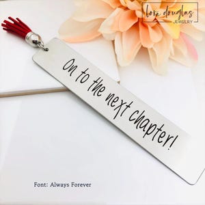 May include: A rectangular silver-toned metal bookmark featuring the black text On to the next chapter! in a script font. The bookmark includes a small silver metal loop at the top attached to a short, vibrant red fabric tassel.