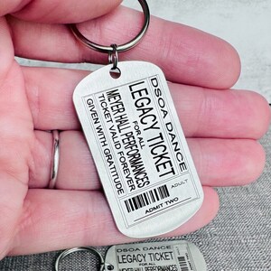 K&ouml;nnte beinhalten: Silberner Schl&uuml;sselanh&auml;nger mit einem Metalletikett, auf dem "DSOA Dance Legacy Ticket for all Meyer Hall Performances Ticket Valid Forever Given with Gratitude" und "Adult Admit Two" steht.