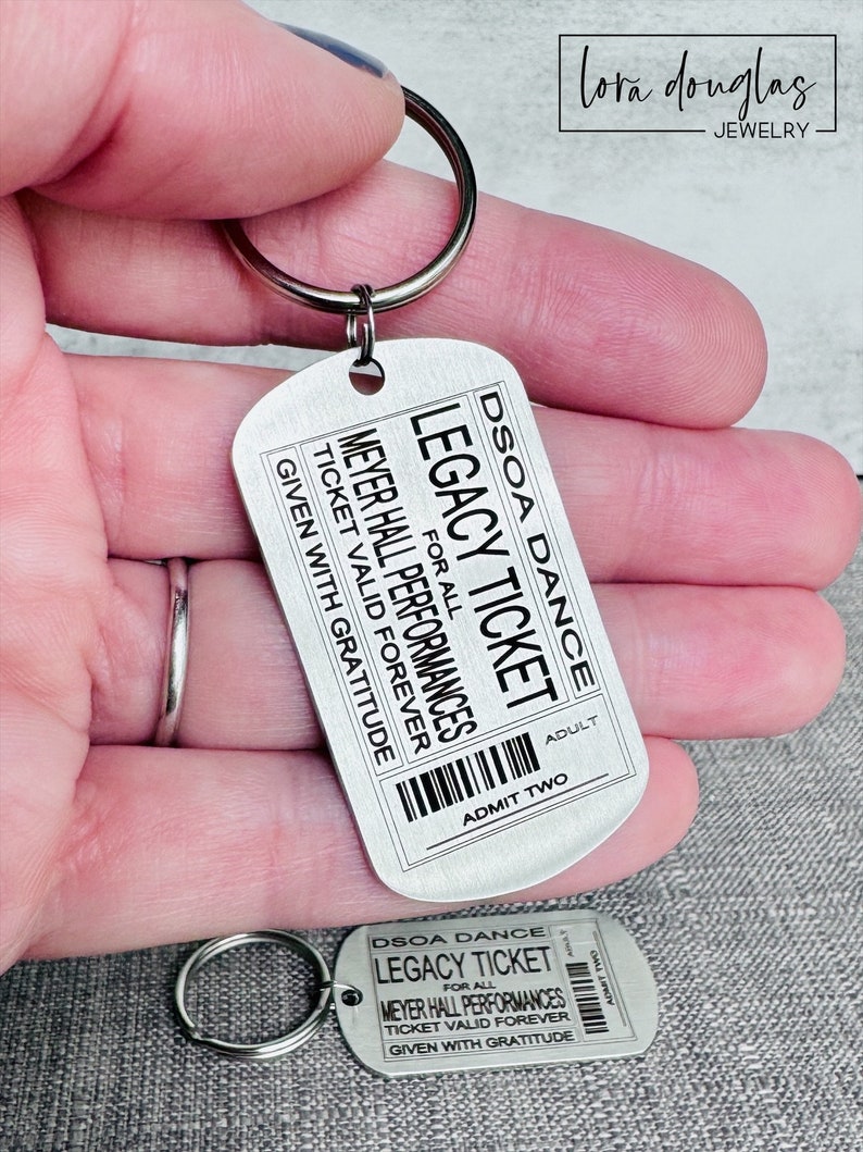 K&ouml;nnte beinhalten: Silberner Schl&uuml;sselanh&auml;nger mit einem Metalletikett, auf dem "DSOA Dance Legacy Ticket for all Meyer Hall Performances Ticket Valid Forever Given with Gratitude" und "Adult Admit Two" steht.