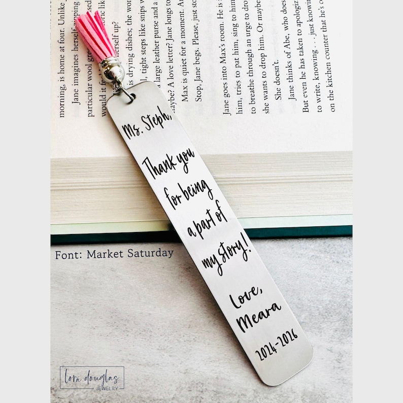 May include: A rectangular silver-toned metal bookmark featuring a pink tassel at the top. The bookmark is engraved with the black text: Ms. Steph, Thank you for being a part of my story! Love, Meara 2024-2026. The metal surface has a smooth, brushed finish.