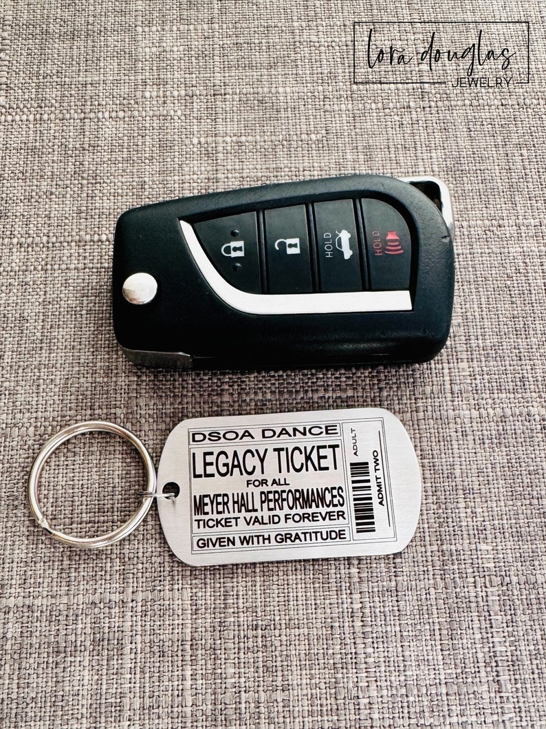 K&ouml;nnte beinhalten: Ein silberner Schl&uuml;sselanh&auml;nger aus Metall mit einem Schl&uuml;sselring und einem Anh&auml;nger, auf dem "DSOA DANCE LEGACY TICKET FOR ALL MEYER HALL PERFORMANCES TICKET VALID FOREVER GIVEN WITH GRATITUDE" und "ADMIT TWO" auf der rechten Seite steht.