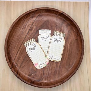 Op de afbeelding: Een rond, donkerbruin houten dienblad met drie overlappende papieren uitsparingen in de vorm van weckpotten. Elke pot heeft het woord Prayer bovenaan, horizontale lijnen om op te schrijven en het woord Amen onderaan gedrukt.