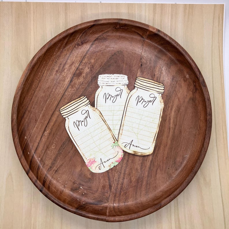 Op de afbeelding: Een rond, donkerbruin houten dienblad met drie overlappende papieren uitsparingen in de vorm van weckpotten. Elke pot heeft het woord Prayer bovenaan, horizontale lijnen om op te schrijven en het woord Amen onderaan gedrukt.