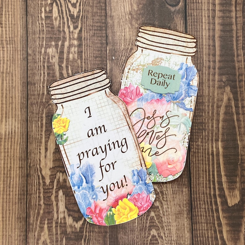 Op de afbeelding: Twee uitsparingen in de vorm van een mason jar met bloemmotieven en tekst. De ene pot toont de woorden I am praying for you! en de andere de tekst Repeat Daily en Jesus loves me, beide versierd met kleurrijke gele, roze en blauwe bloemen.