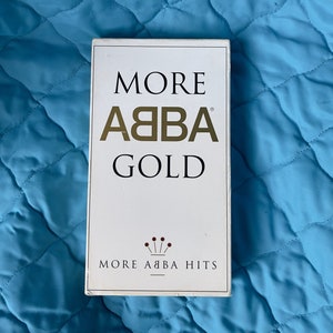 Pode incluir: Uma caixa de fita cassete branca com letras douradas que lê "More ABBA Gold More ABBA Hits".