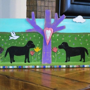 May include: A hand-painted wooden art piece depicting two black dogs in a field of green grass with pink and yellow flowers. A purple tree with a pink heart is in the center of the piece. The piece has a mosaic tile border.