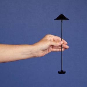 Puede incluir: Una pequeña lámpara negra de forma triangular, con un tallo largo y delgado y una base circular. La lámpara se sostiene sobre un fondo azul. La altura de la lámpara es de aproximadamente 20 cm.