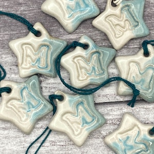May include: Eight ceramic star ornaments with a blue and white glaze. Each star has a raised letter "M" in the centre. The stars are strung with twine.
