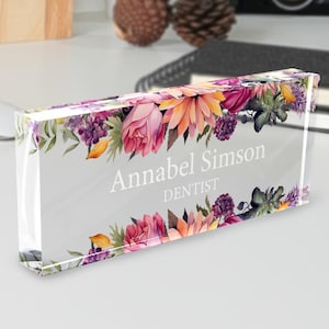 May include: A clear acrylic desk nameplate with a floral design. The nameplate features the name "Annabel Simson" and the title "DENTIST" in white text. The floral border includes pink, orange, and purple flowers.