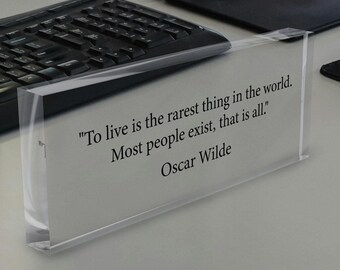 May include: Clear acrylic block with the quote "To live is the rarest thing in the world. Most people exist, that is all." by Oscar Wilde. The block is on a white surface, near a keyboard and a black tablet.