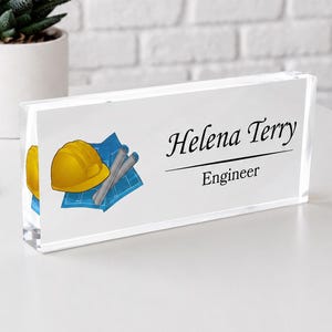 May include: A rectangular clear acrylic desk name plate featuring the printed name Helena Terry and the title Engineer. The design includes a graphic of a yellow hard hat resting on a blue architectural blueprint with rolled documents.