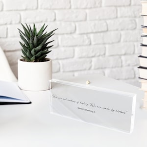 May include: A clear acrylic block with the quote "We are not makers of history. We are made by history." by Martin Luther King Jr. The block sits on a white surface with a small potted succulent and a stack of books in the background.
