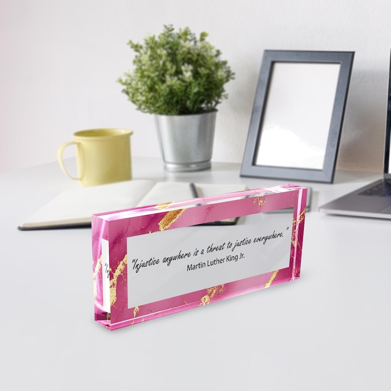 May include: A pink and gold decorative block with the quote "Injustice anywhere is a threat to justice everywhere." by Martin Luther King Jr. The block is rectangular and sits on a white surface.