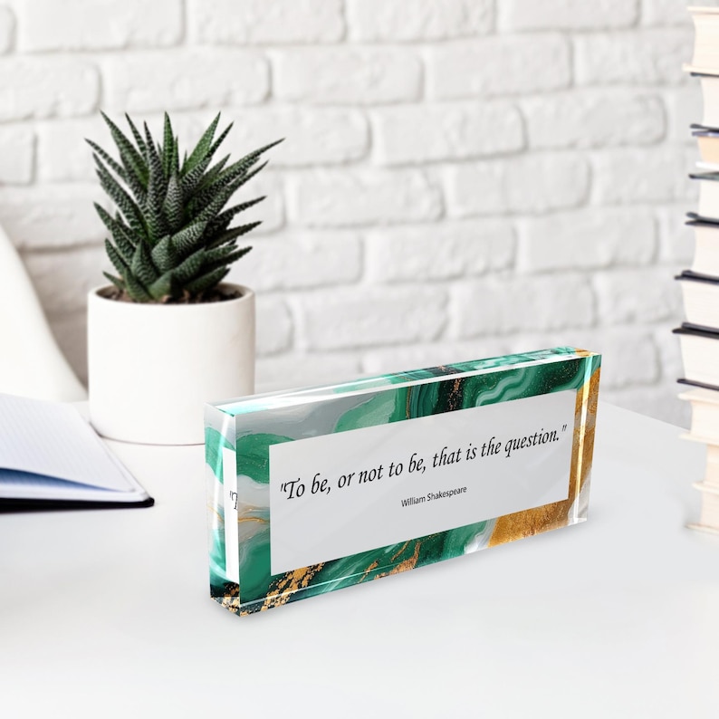 May include: A rectangular desk block with a quote from William Shakespeare, "To be, or not to be, that is the question." The block has a green and gold marbled design. A small succulent plant in a white pot is in the background.