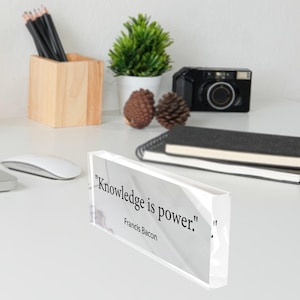 May include: A clear acrylic block with the quote "Knowledge is power." by Francis Bacon. The block is on a white surface, with a wooden pencil holder, a small plant, and a camera in the background.